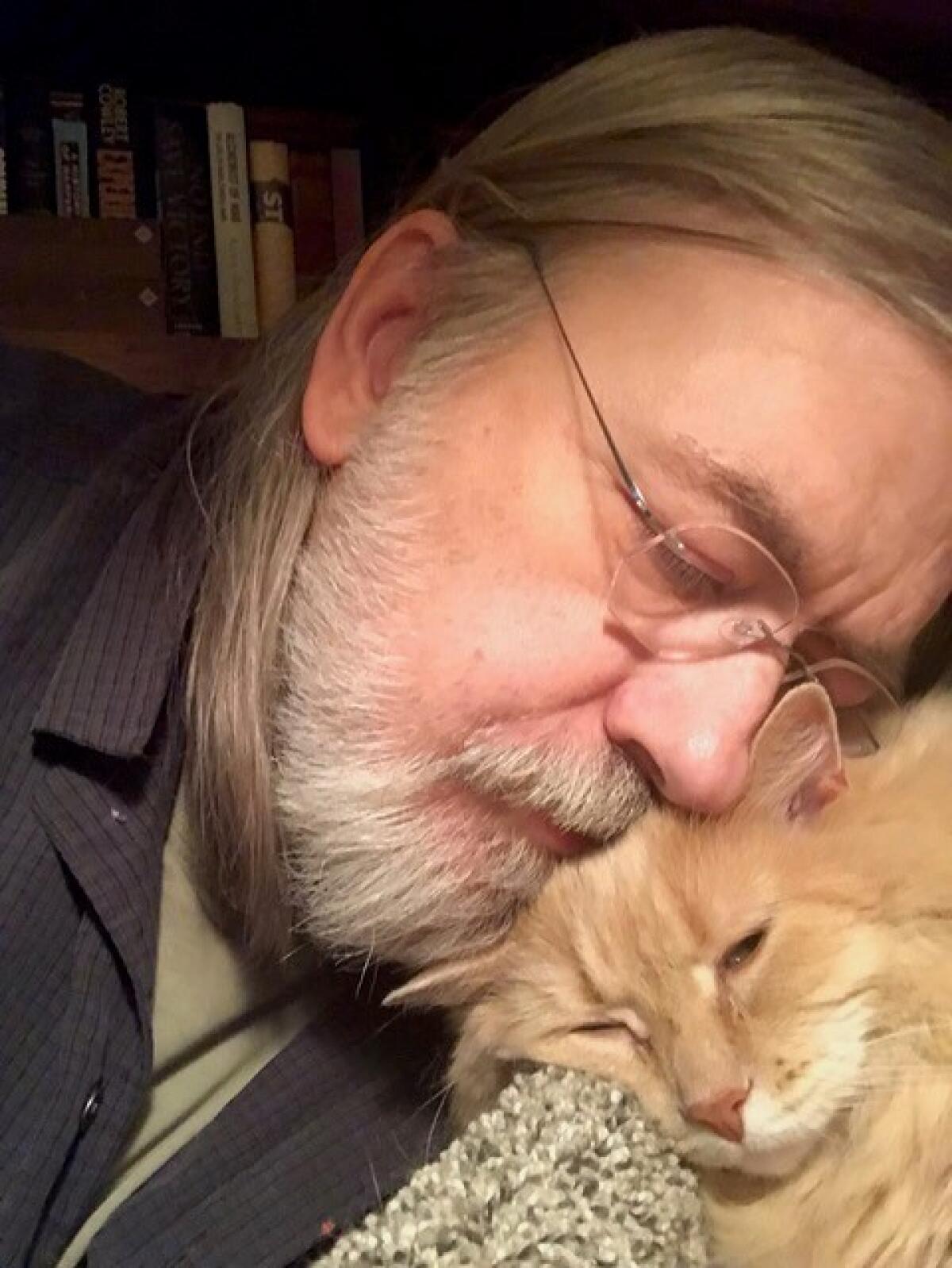 Caleb Carr, with glasses and a white beard, nestles his face on his cat's head.