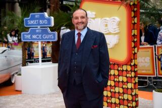 Joel Silver smiles and poses with his hands in his pockets while wearing a blue suit in front of a colorful sign.
