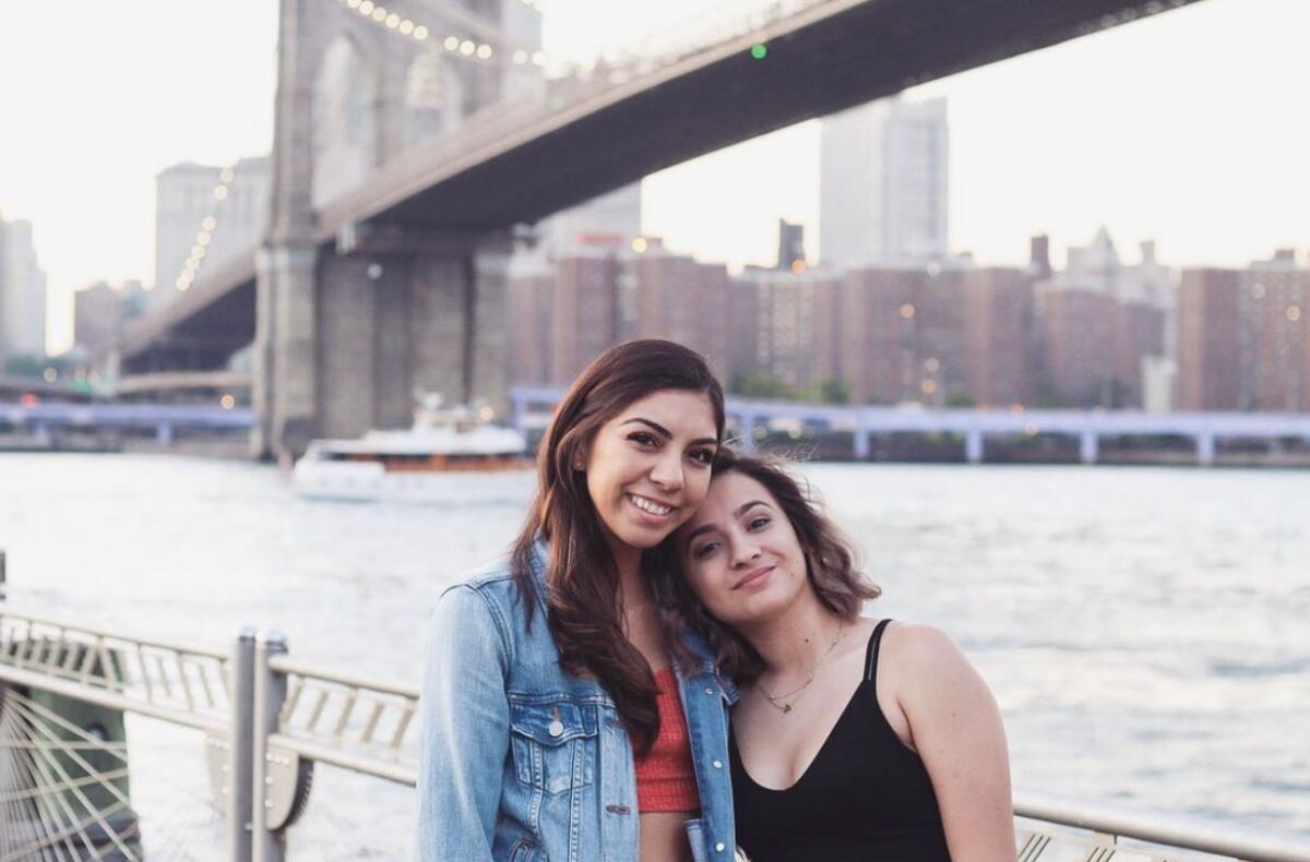 Two young women, in a head-and-shoulders frame, press their heads together, smiling for a photo in front of Brooklyn Bridge