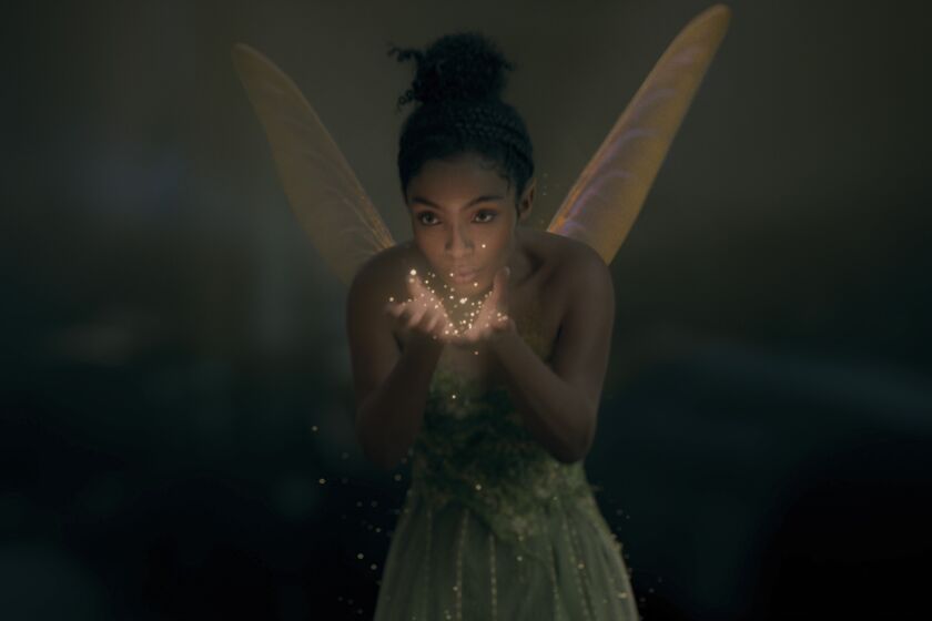 A woman with black hair in a bun and fairy wings wearing a green dress and blowing sparkles
