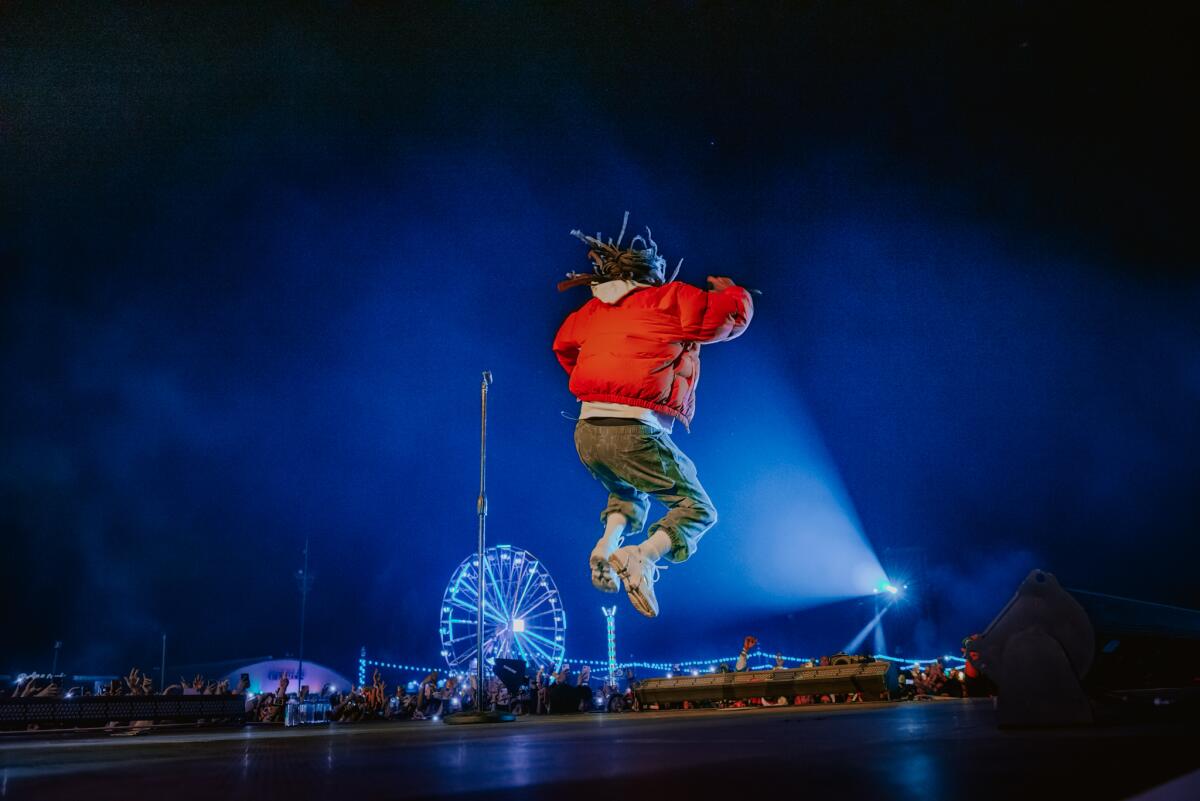 A rapper leaps into the air onstage at a music festival, with a Ferris wheel in background