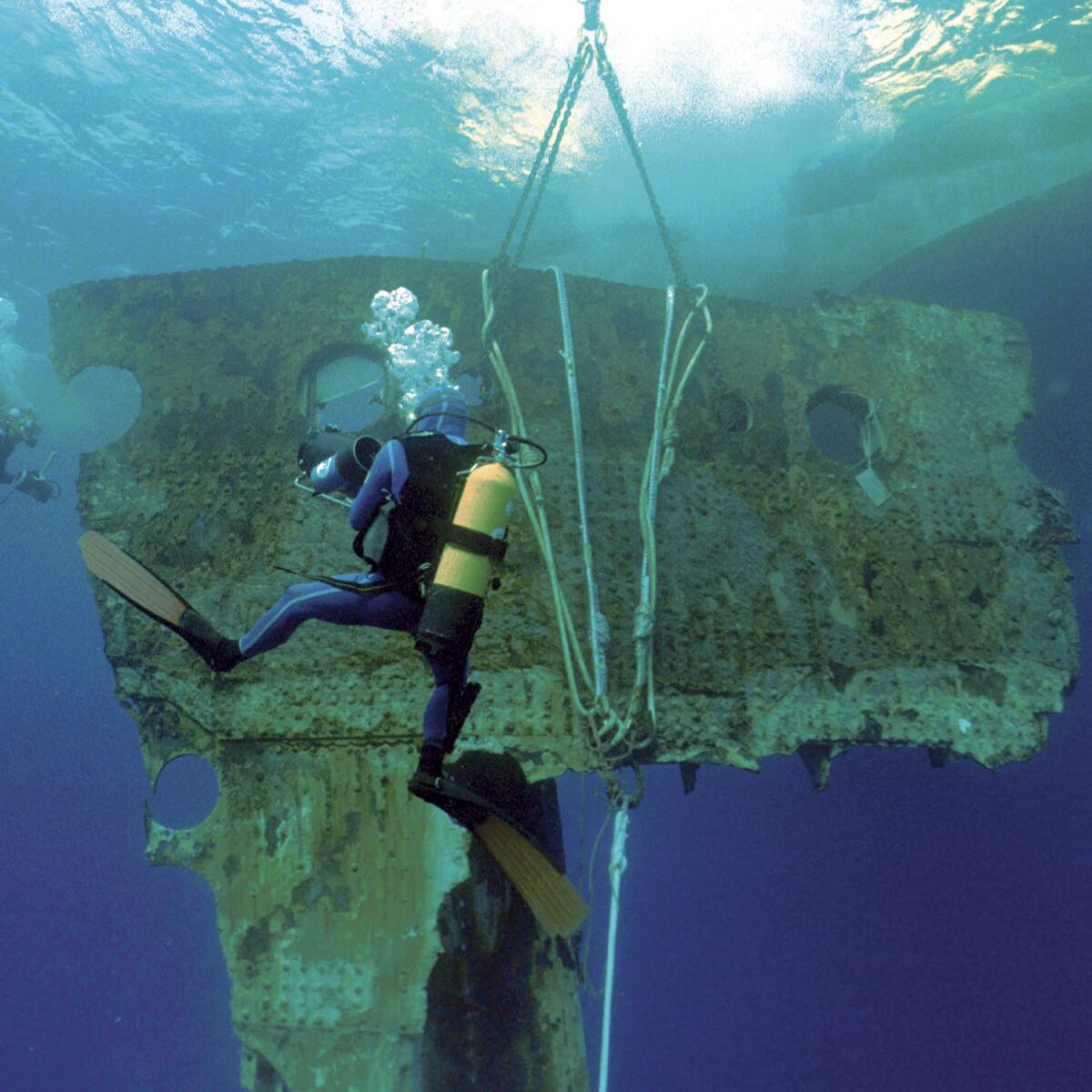 An underwater scene of a diver with an oxygen tank swimming vertically in front of a large metal piece of shipwreck