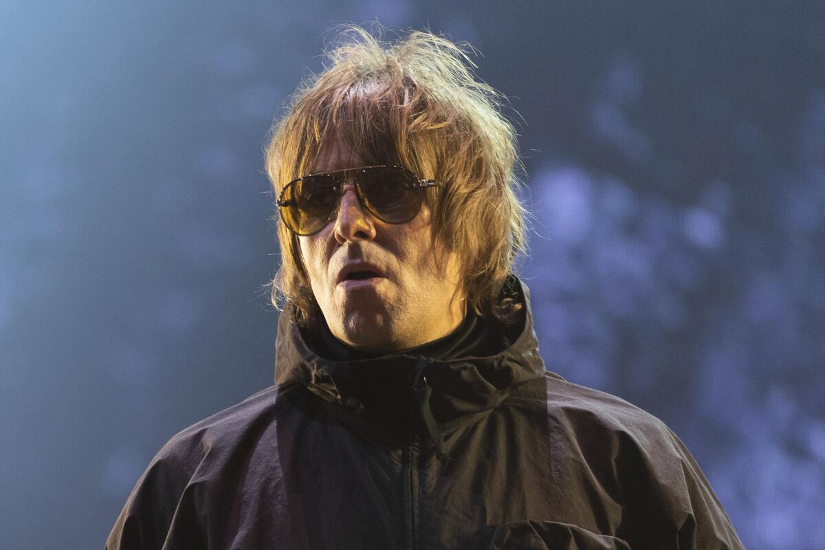 Liam Gallagher performing in a black anorak and sunglasses with tousled hair and a weird facial expression