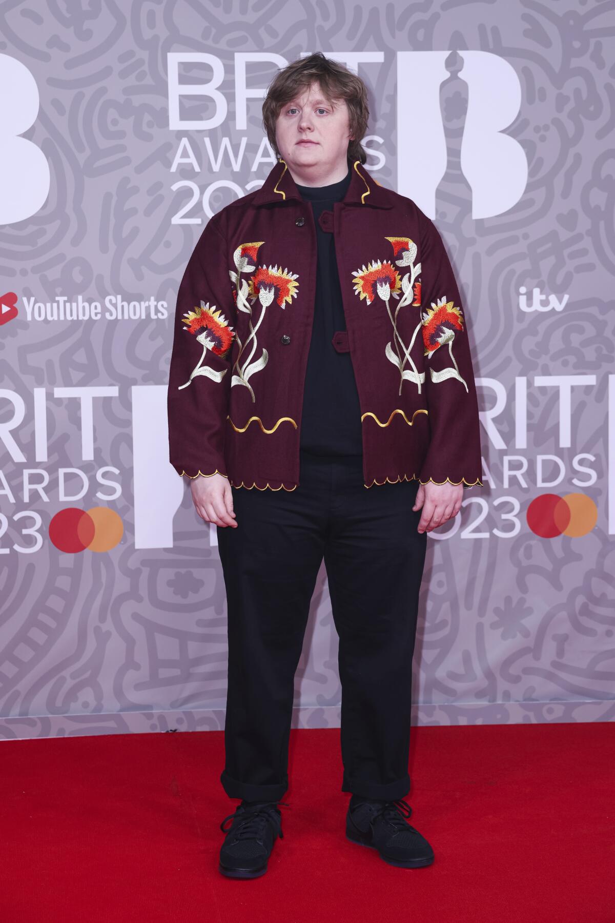 Lewis Capaldi poses for photographers with arms to the side, wearing a maroon coat with flower designs and black pants