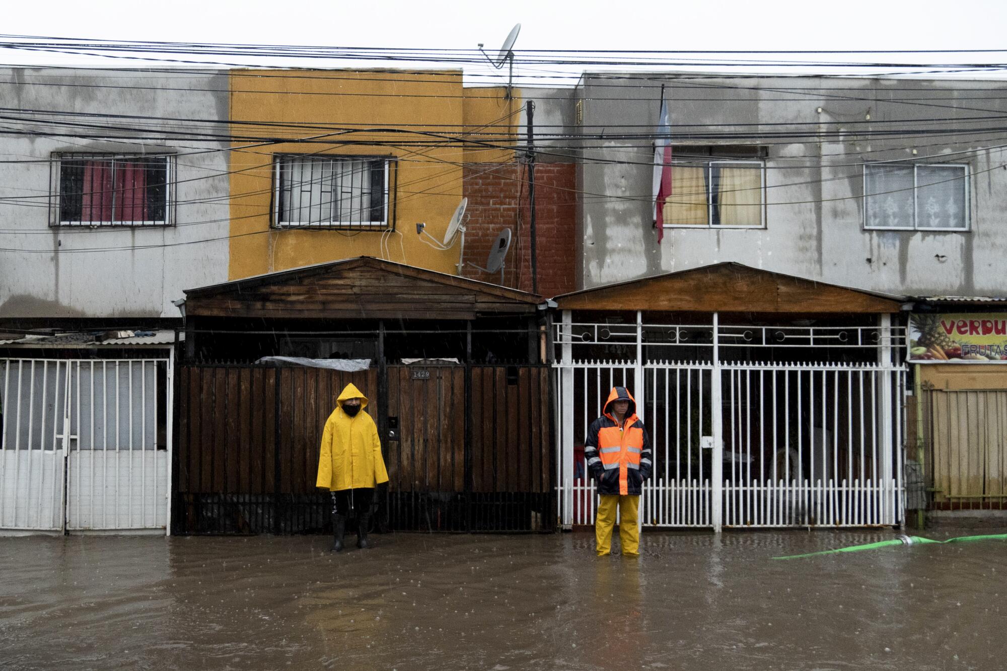 Two people, one in a yellow rain jacket, stand on a flooded street outside homes 