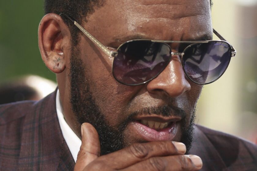 A tight closeup on the face of a man in a suit and sunglasses holding his hand near his bearded chin