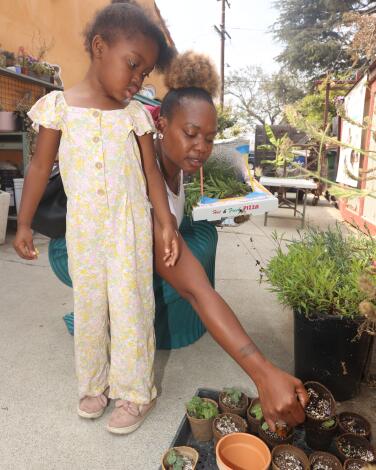 A little girl stands next to a woman kneeling to tend to small potted plants