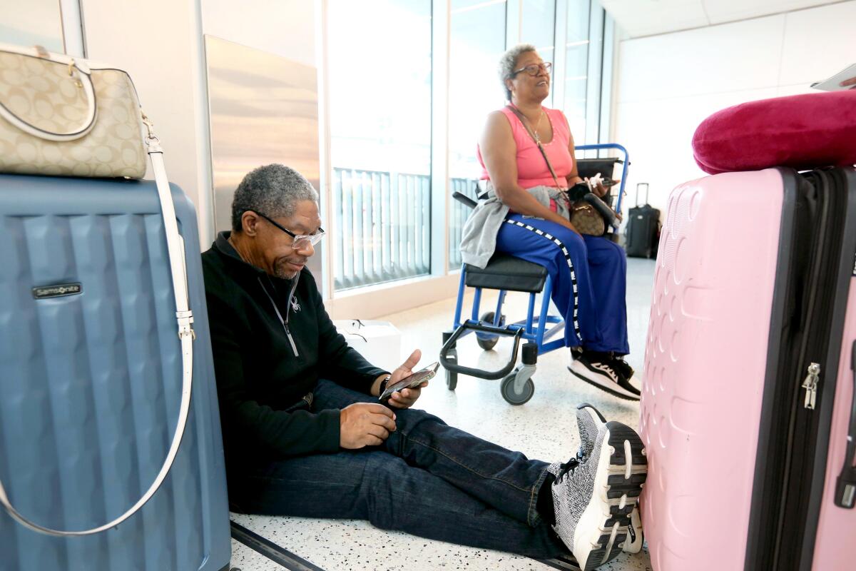 A man sits on the ground between two suitcases and a woman sits in an airport wheelchair nearby.