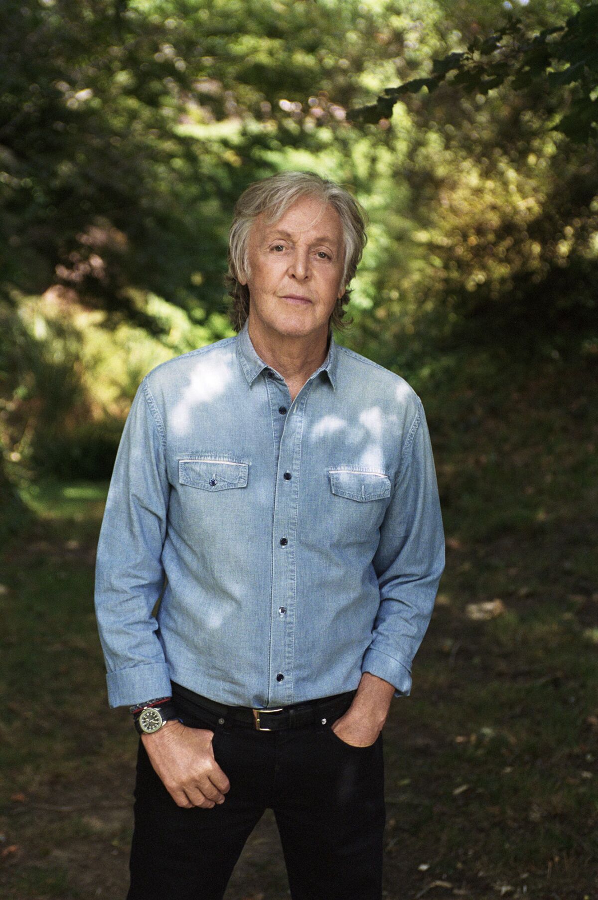 Paul McCartney stands outdoors, hands in pockets, in a blue denim shirt and dark pants.