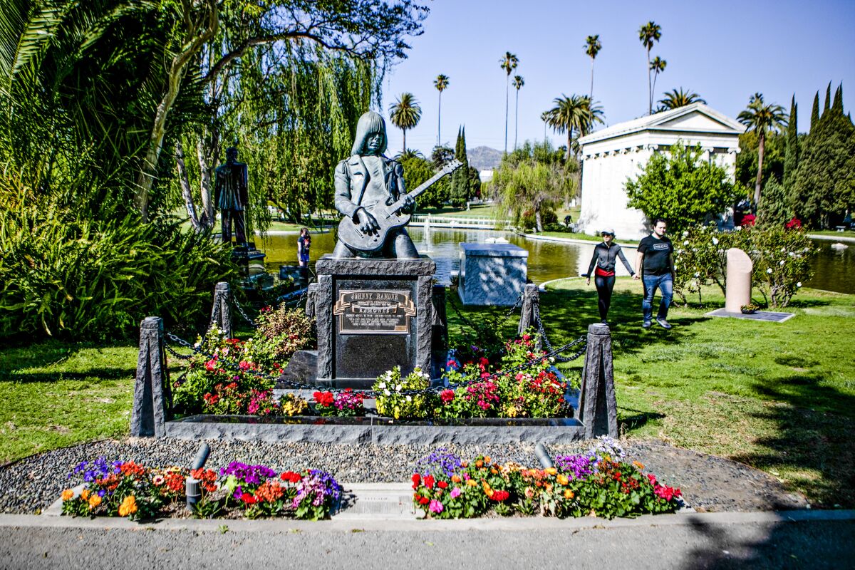 A life-size guitar-playing man marks a grave at the Hollywood Forever Cemetery, with a white mausoleum in the distance.