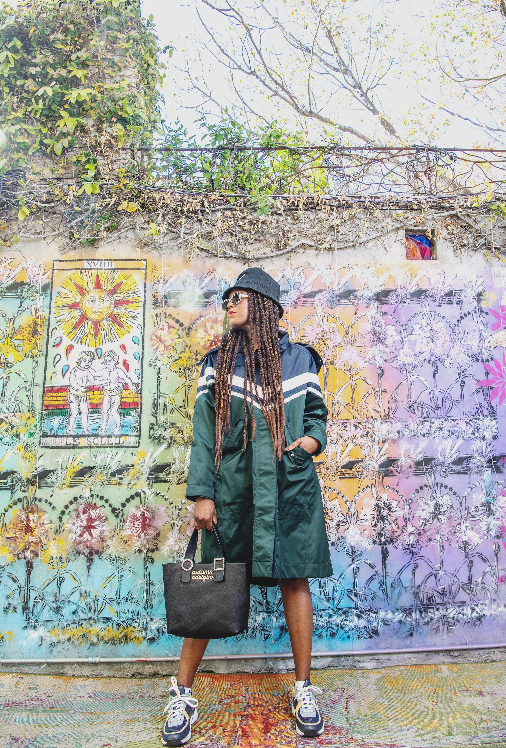 Autumn Adeigbo in a long coat and hat with a purse in front of a wall with a repeating colorful image