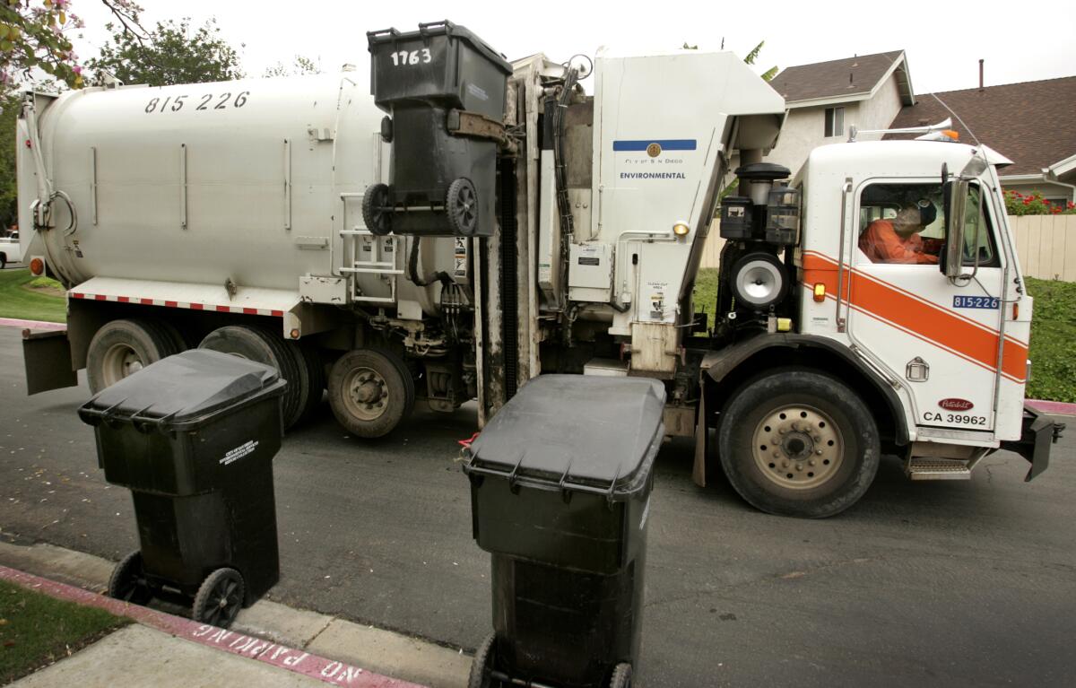 A garbage truck driver on a residential street looks up at a trash bin being lifted to be emptied into his truck