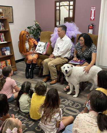 Children sit on the floor while adults read to them and a dog looks on in a bookstore.