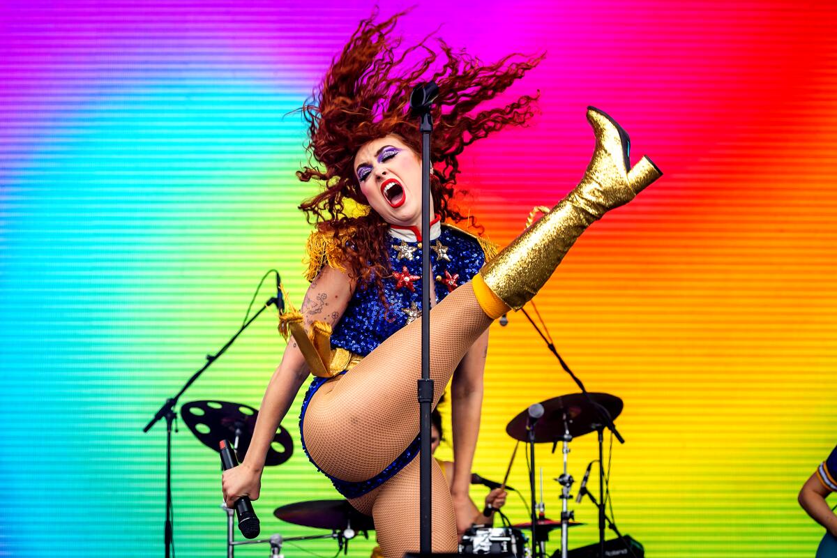 A woman with whipping hair kicks up her booted leg as she sings into a microphone behind a rainbow-colored backdrop.