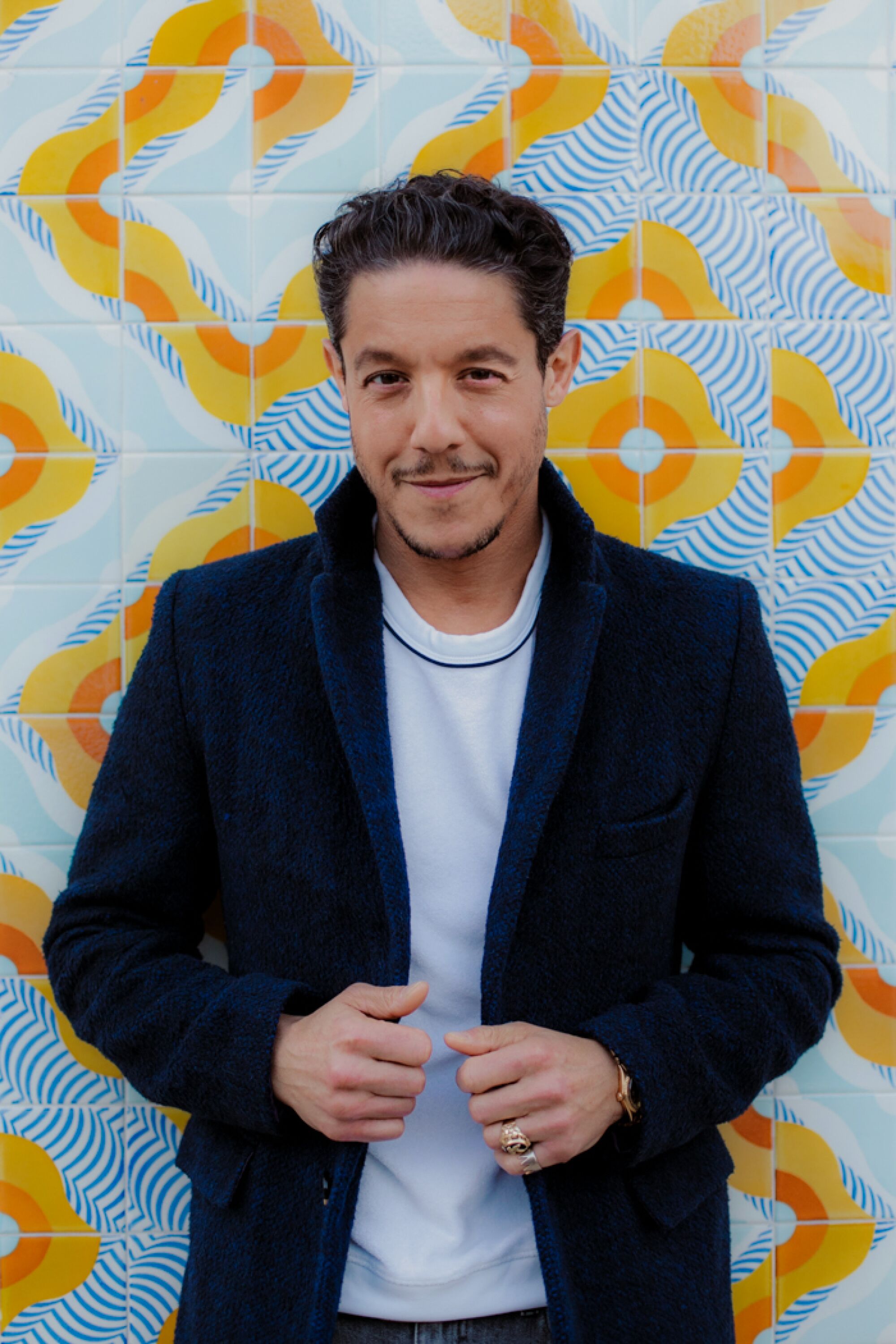 A man in a white shirt and yellow jacket stands in front of a patterned wall.