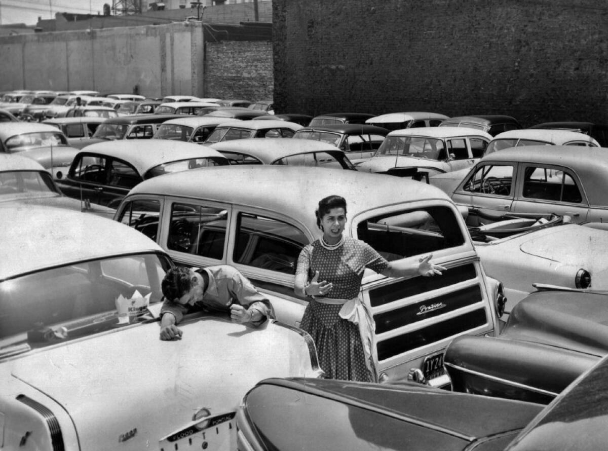 A woman stands and a man hangs over the trunk of a car in a bumper-to-bumper jammed parking lot.