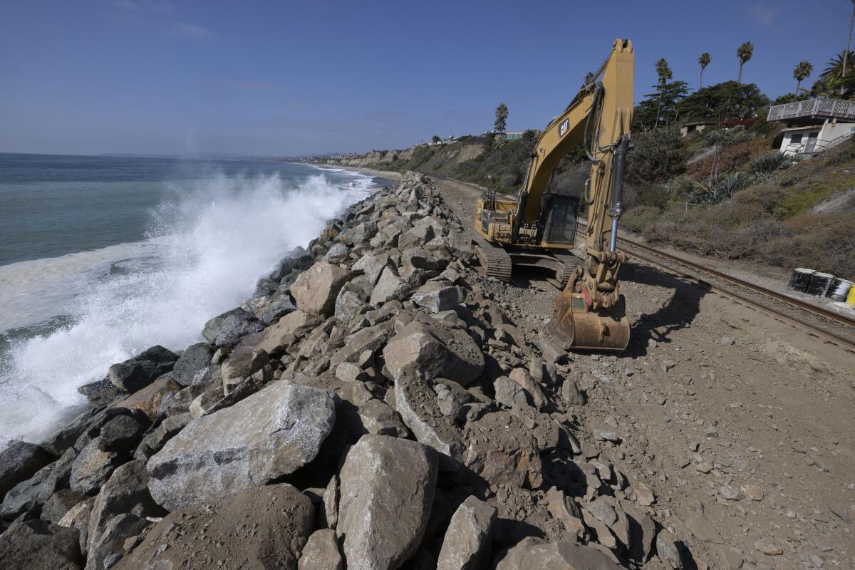 An excavator sits parked next to railroad tracks separated from the ocean by boulders
