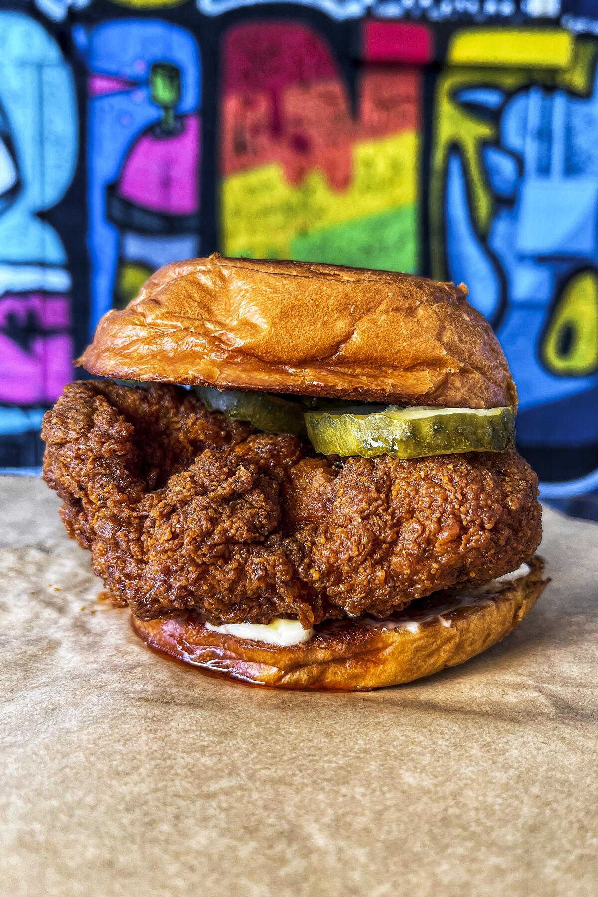 A photo of a pickle-topped fried chicken sandwich against a colorful wall at Bellflower's SteelCraft food hall.