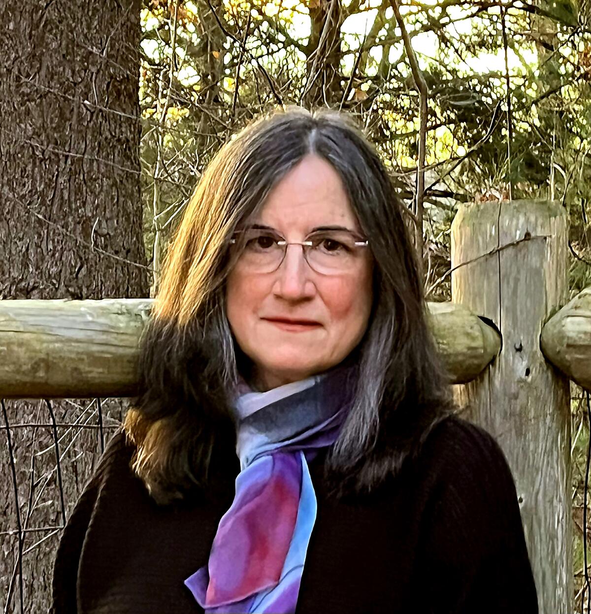 A woman wearing glasses and a colorful scarf looks at the camera, outside in a woodsy area.