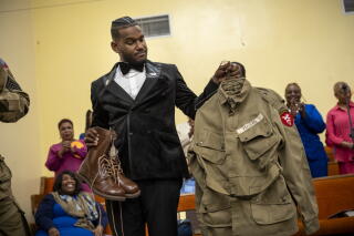 A man in a suit holds a pair of combat boots and a paratrooper uniform.