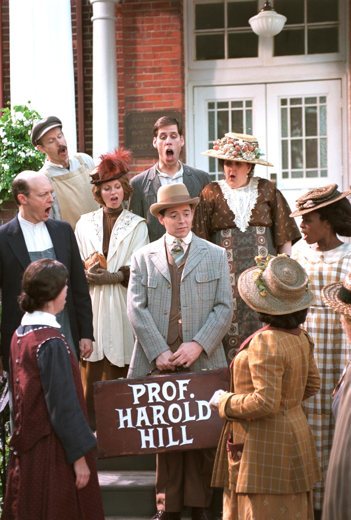 Matthew Broderick, in costume and holding a suitcase labeled "Prof. Harold Hill," is surrounded by singing men and women.