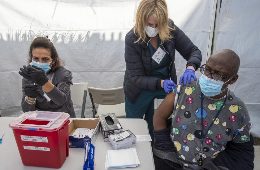At a vaccination site, a health worker vaccinates a man while another health worker prepares a syringe.