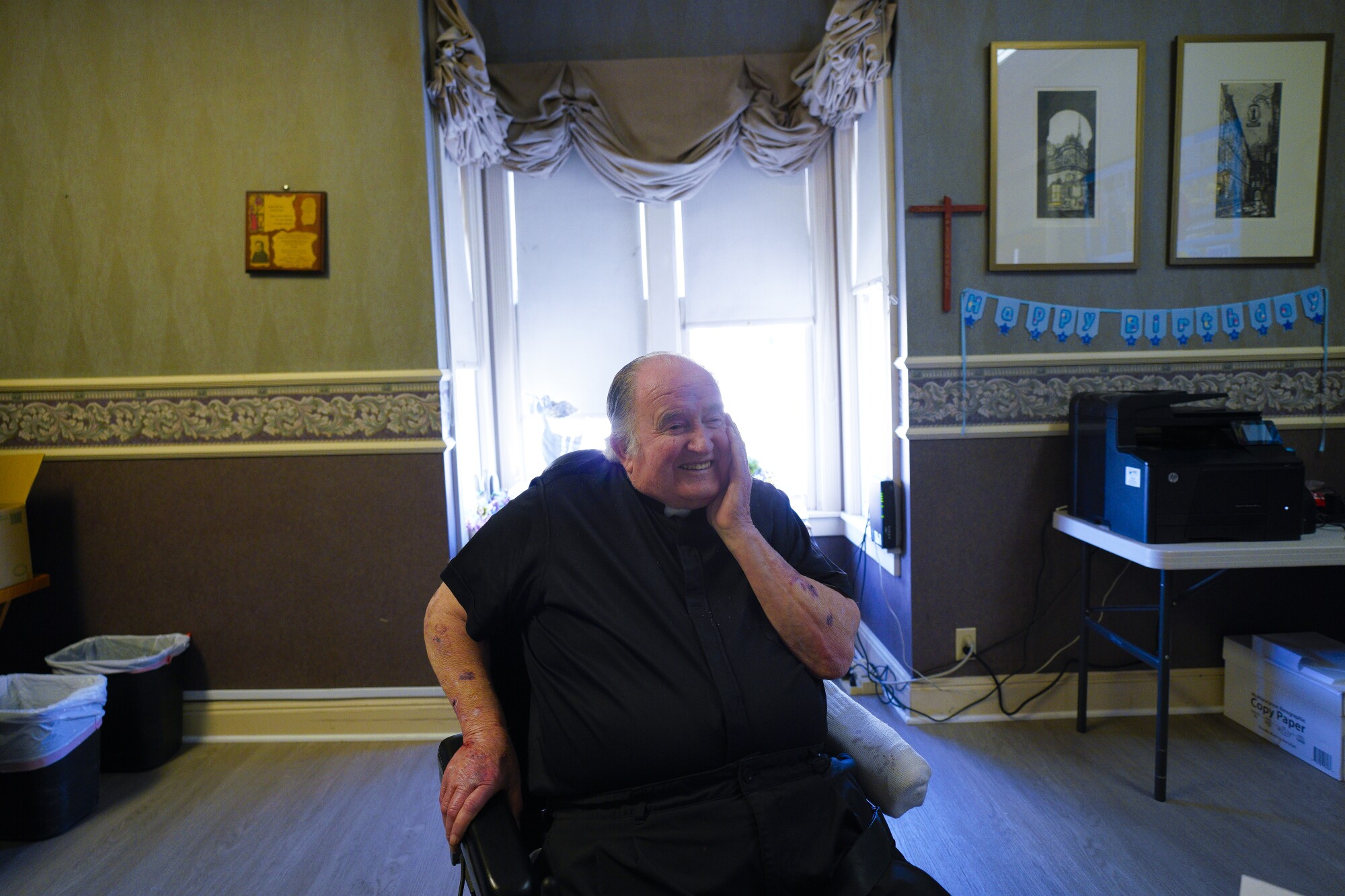 Father Joe Carroll sits in a chair in a room while holding his hand to his face