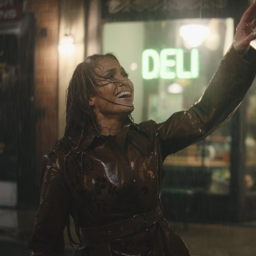 Jennifer Lopez in the rain, outside a deli storefront, waving her left hand and smiling