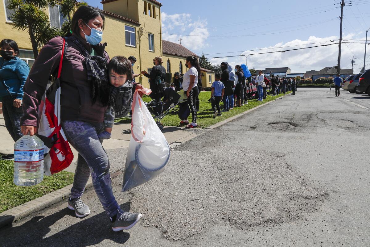 A woman with a toddler strapped to her chest holds a jug of water and a bag 