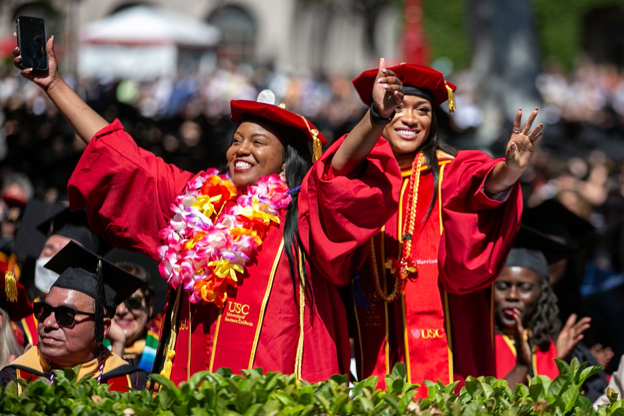 Two USC graduates, both in red robes and one wearing a lei, smile at commencement.
