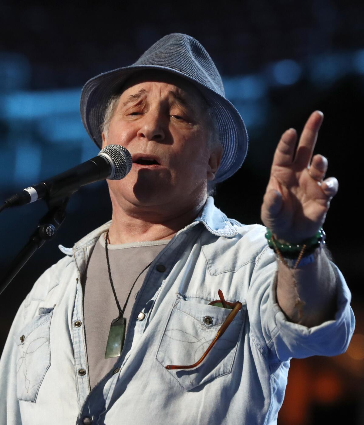 Paul Simon sings on stage with his left hand outstretched while wearing a gray fedora and casual shirt