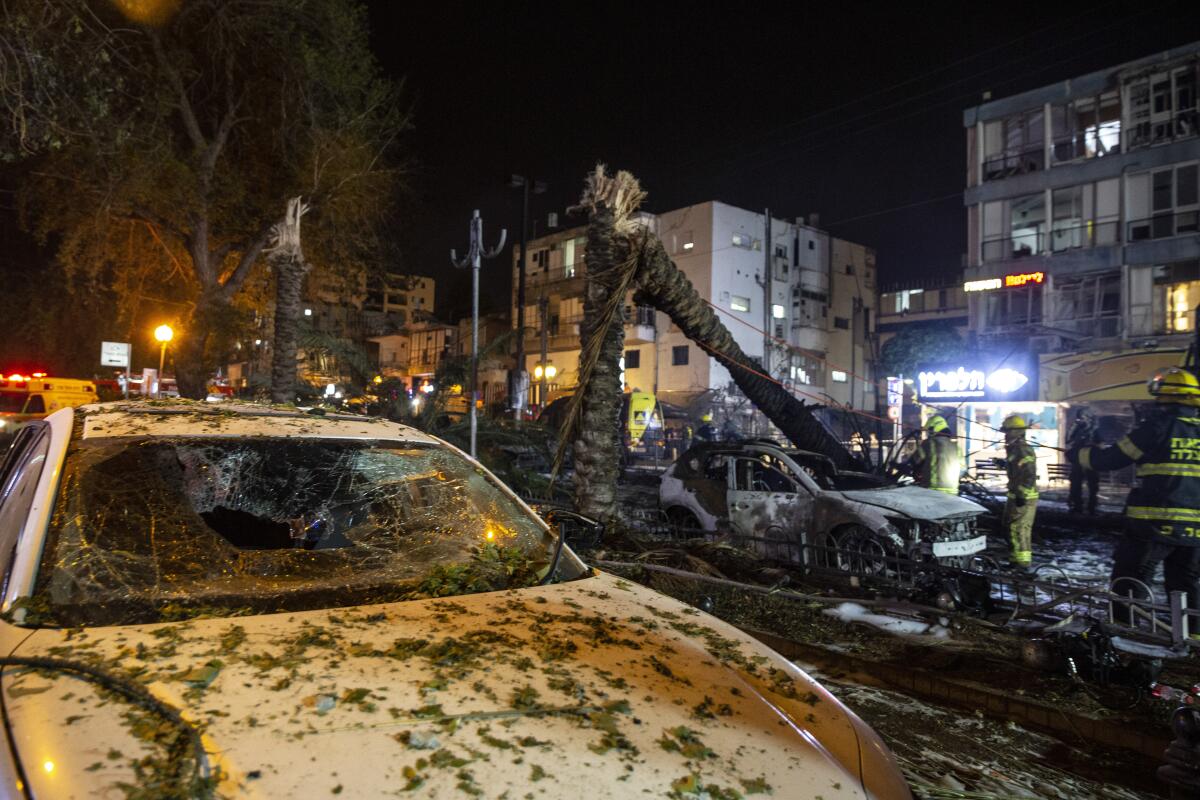 A car with a broken windshield, foreground, near other damaged cars, trees and buildings. 