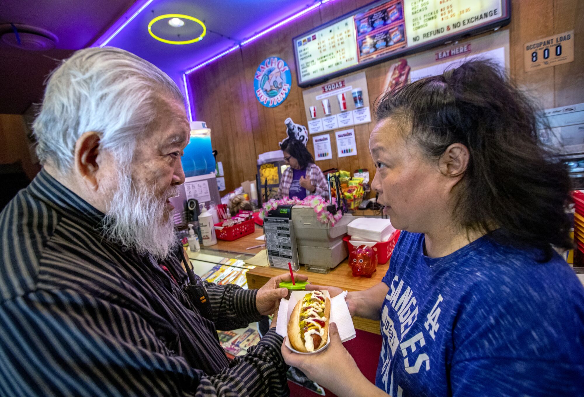 A gray-haired man talks with a woman as she hands him a hotdog inside a cinema foyer