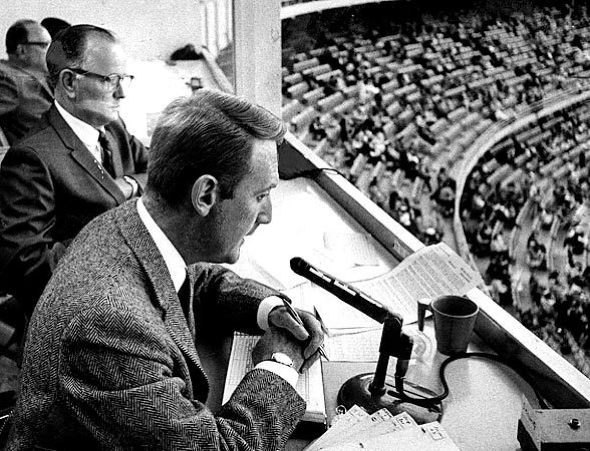 Two men in suits sit in a broadcast booth overlooking a baseball stadium. One speaks into a microphone.