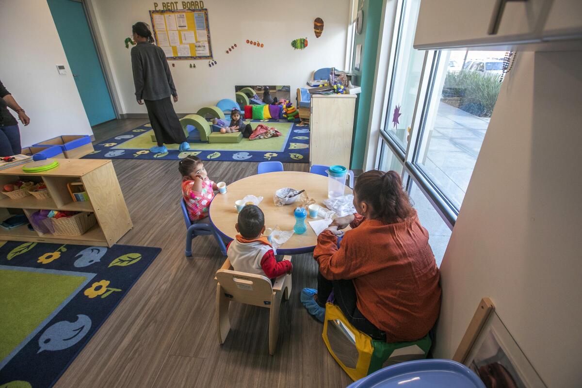 A woman sits at a small table in a child care facility with two toddlers