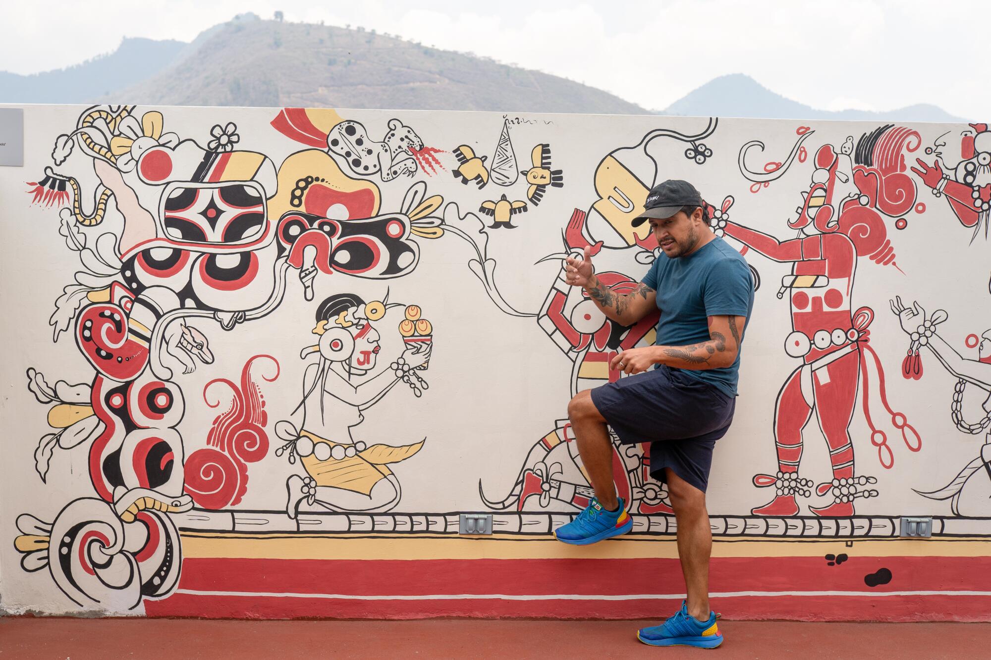 Juan Pablo Romero stands in front of a mural at El Patojismo that resembles a Mayan Codex.