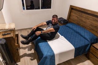 A man with a dark beard, in dark clothes, sits on a bed with blue and white linens