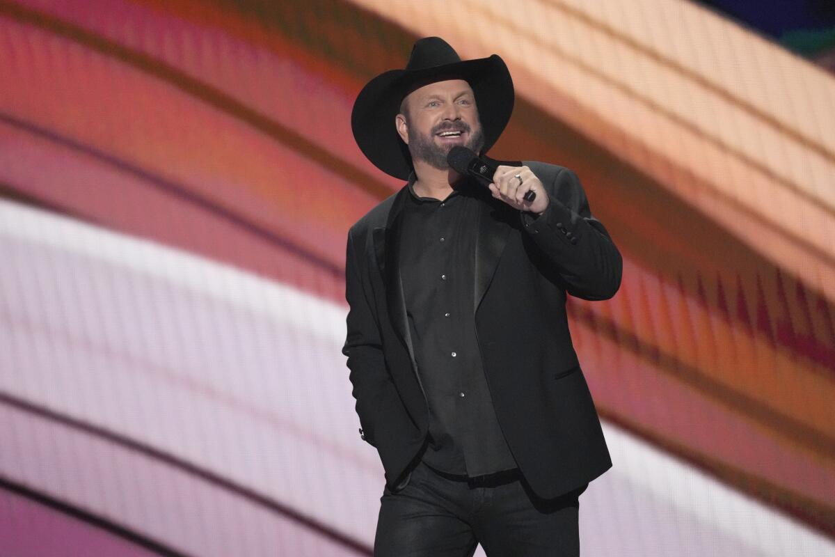 A smiling bearded man in a black outfit and black cowboy hat holds a microphone.