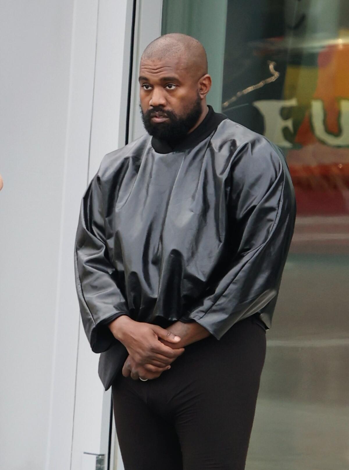 Kanye West poses with a glum look on his face and his hands folded in front of him while wearing a shiny leather coat