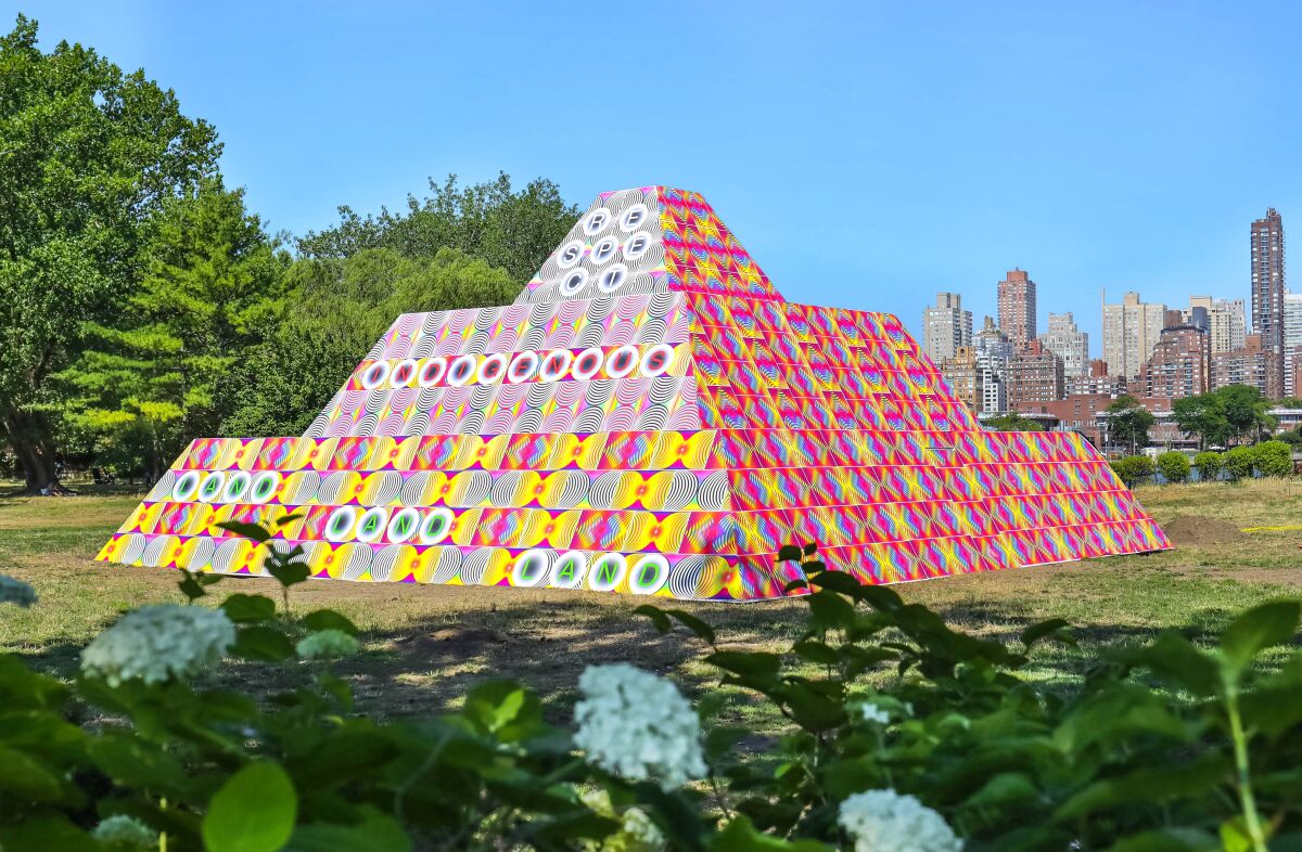 A brilliant pyramid in pink and yellow patterns stands before the New York skyline.