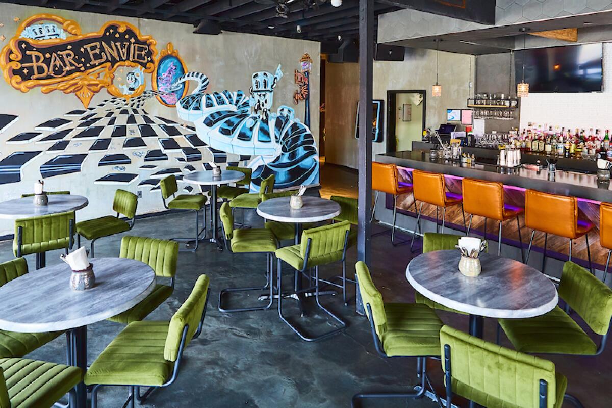 The dining room of Long Beach's Bar Envie, with green chairs at tables, orange chairs at the bar, and a playful mural