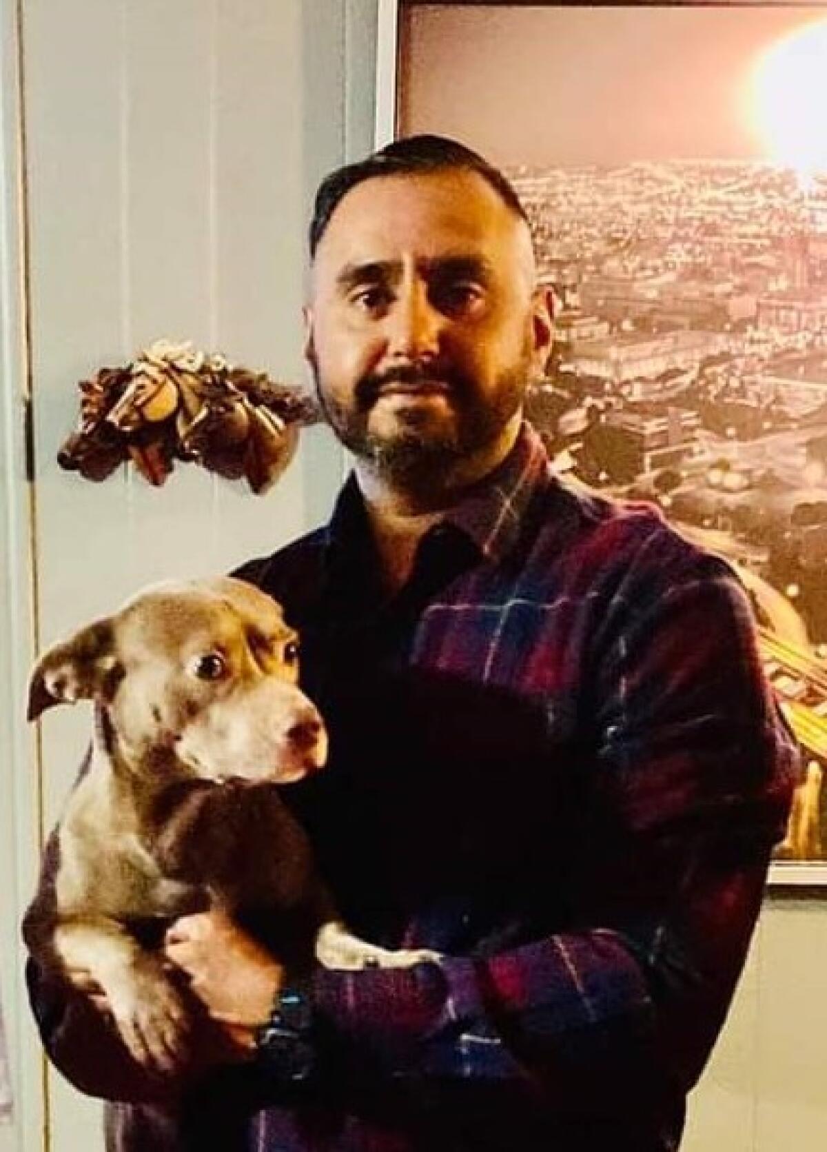 A man with trimmed facial hair poses for a photo while holding a light-brown dog.