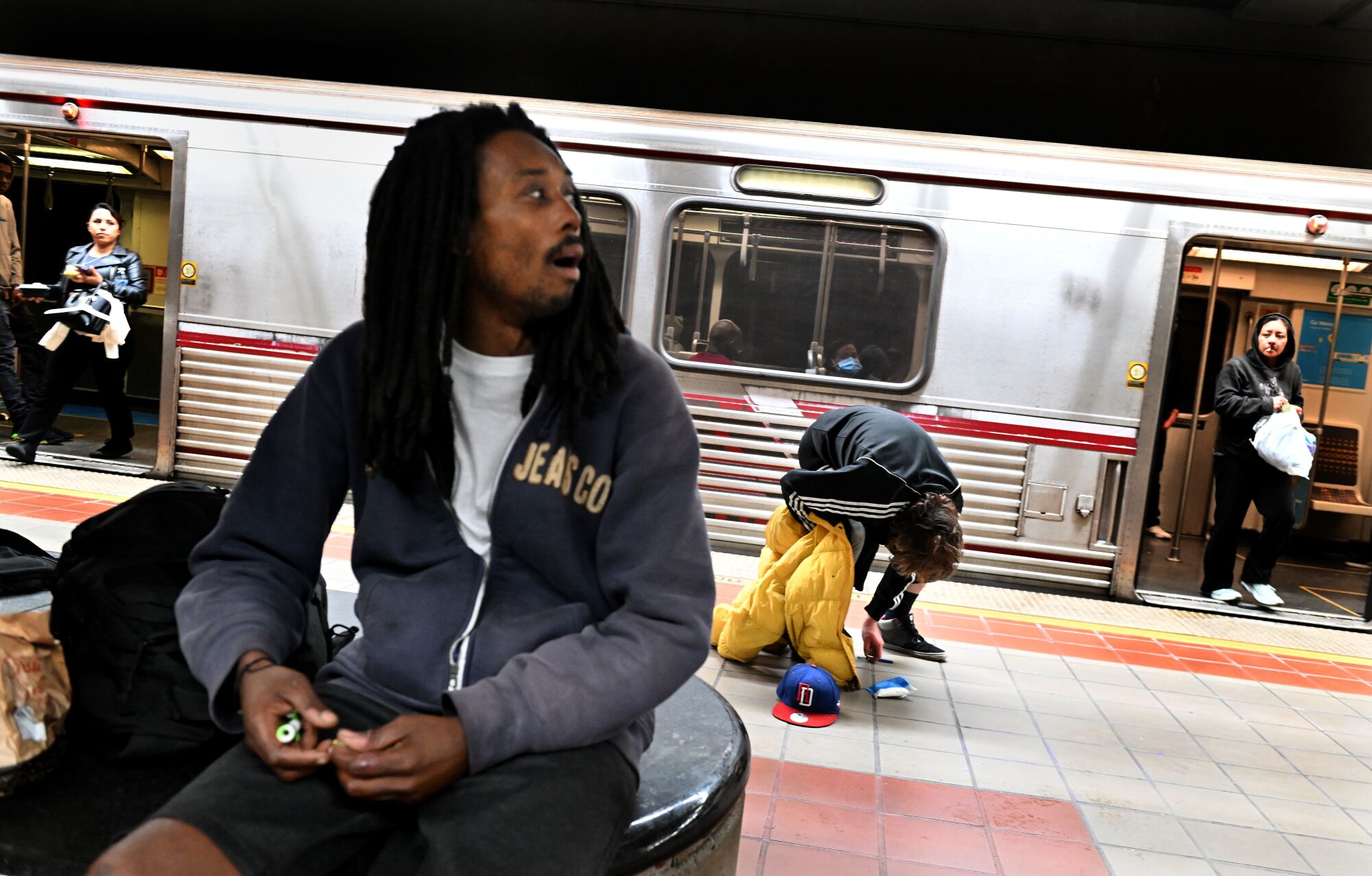 A man bends down and another one sits on a subway bench. A train with doors open is in the background with people exiting.