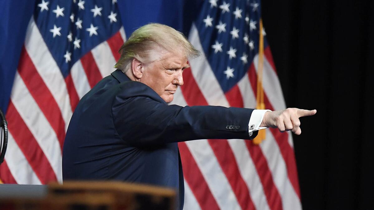 A man with blond hair, in a dark suit, seen from the side, points with two U.S. flags in the background 