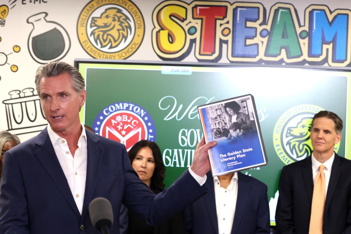 A man in a dark jacket and white shirt holding up a booklet at a school