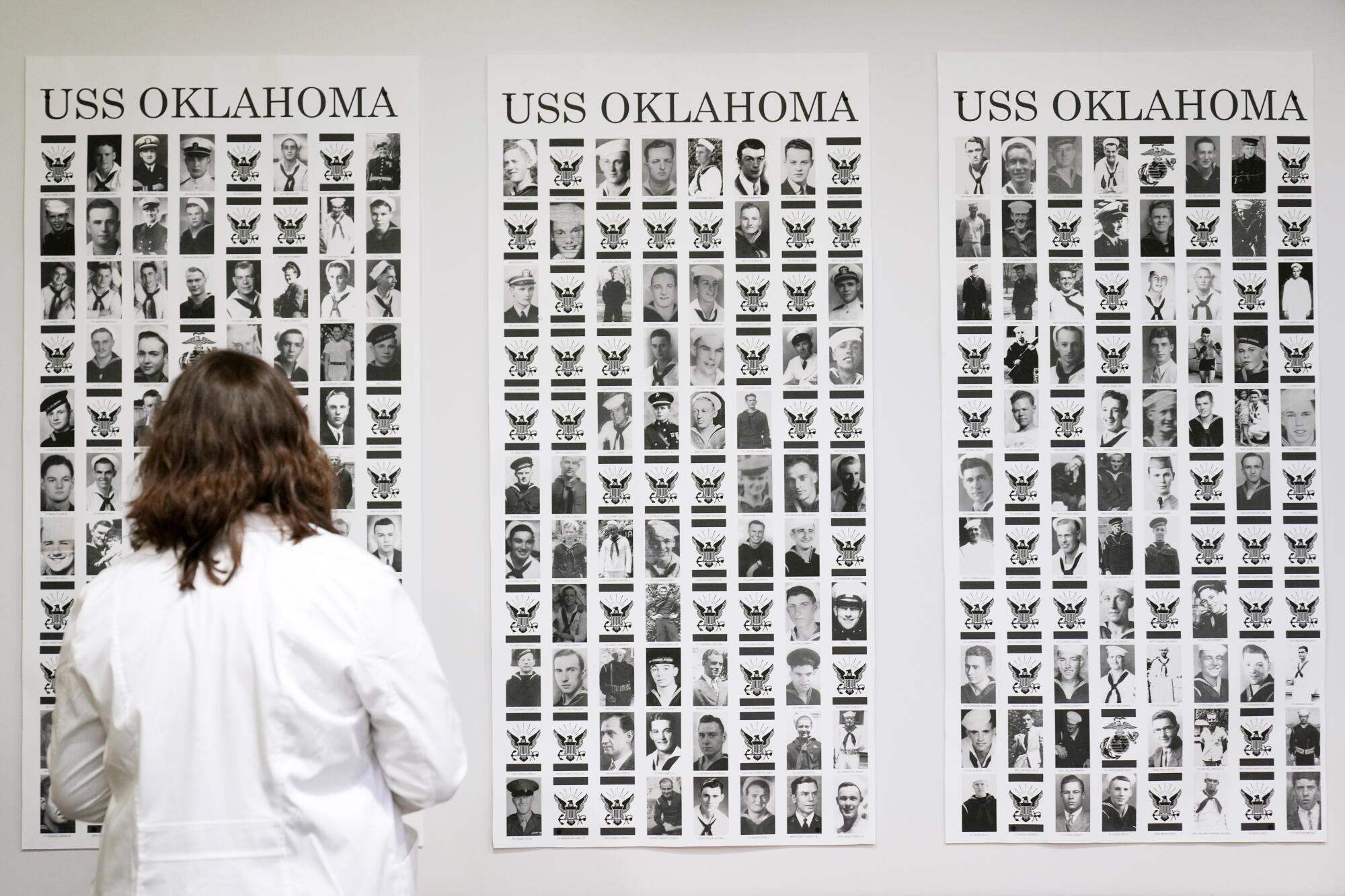 A woman in white, seen from behind, looks at three sets of multiple photos of people, under the label USS Oklahoma