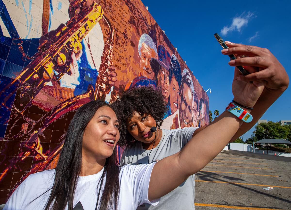 A woman stands in front of an outdoor mural taking a selfie while another woman poses with her