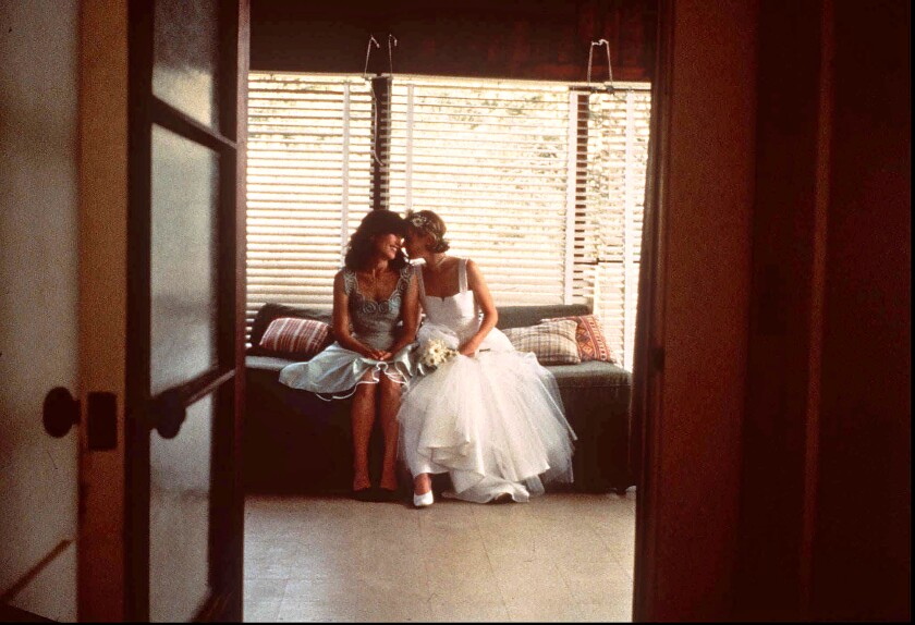 Two women, one in a wedding dress, sit and lean their heads together.