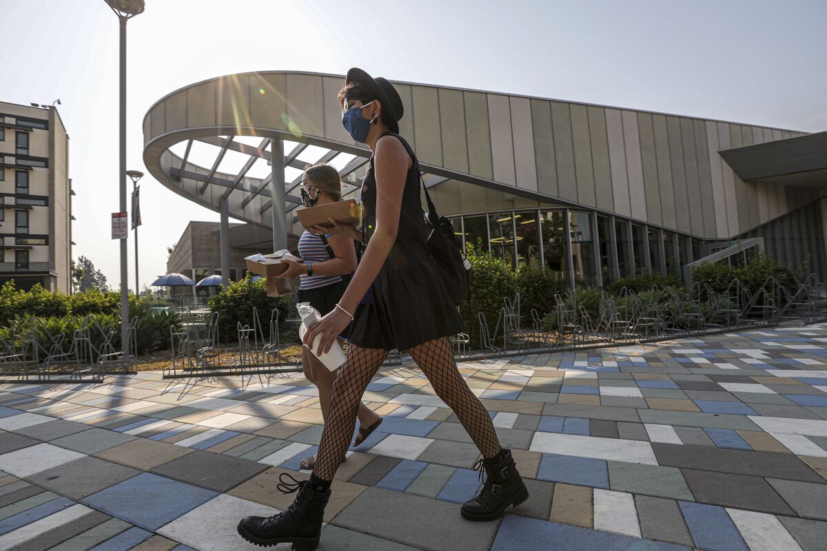 Two women wearing masks walk on a college campus while carrying takeout boxes