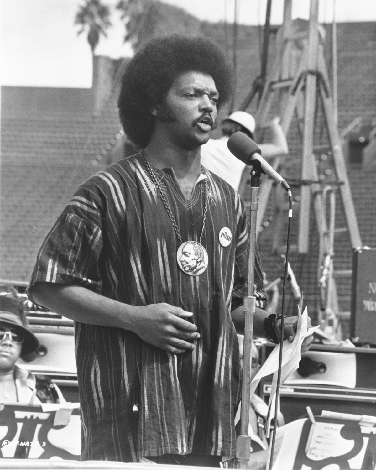 A Black man with an afro, wearing a dashiki and speaking from an outdoor stage
